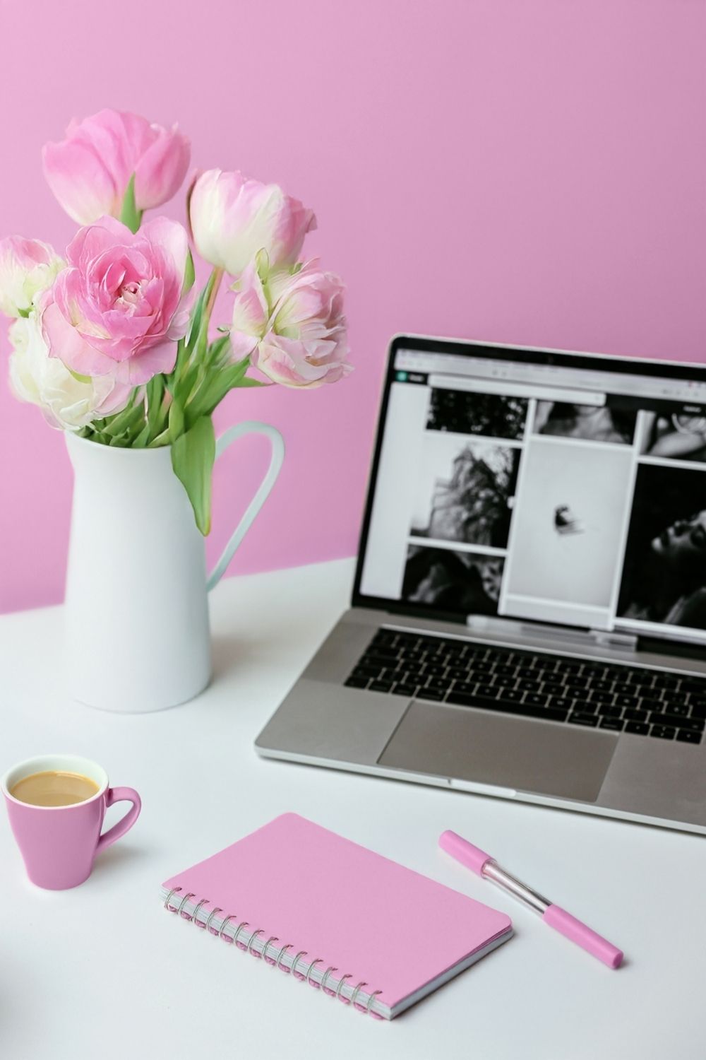 Pink wall with white vase of pink flowers, laptop, pink pen, notebook, a cup on a white desk.