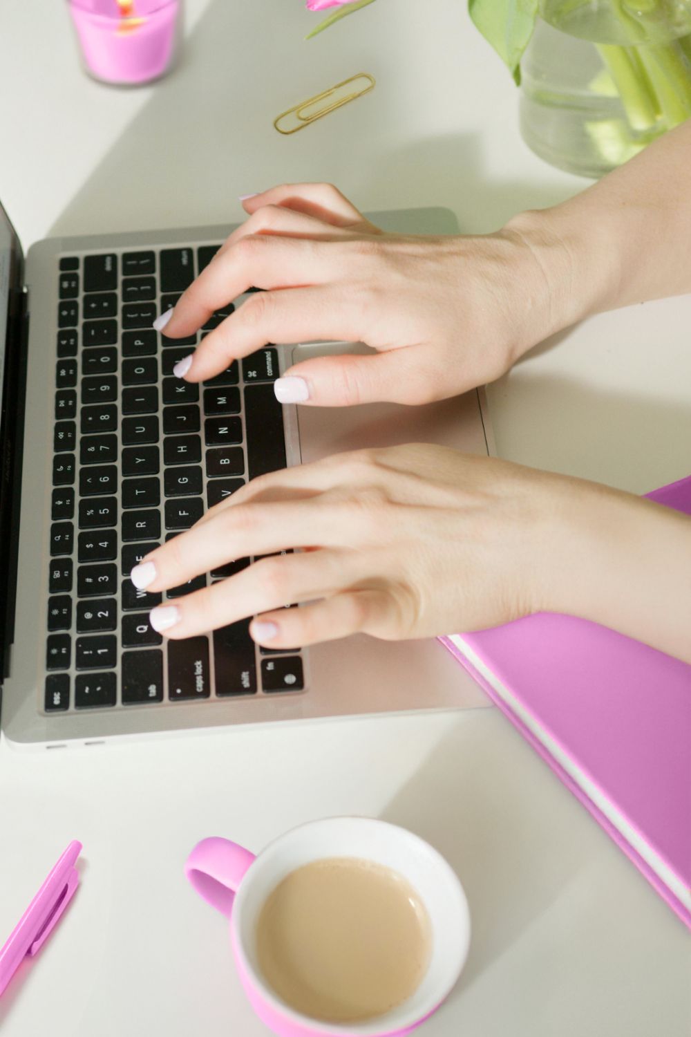 Woman typing on a laptop, surrounded by pink notebooks, coffee, pen, candle, flowers, a gold clip.