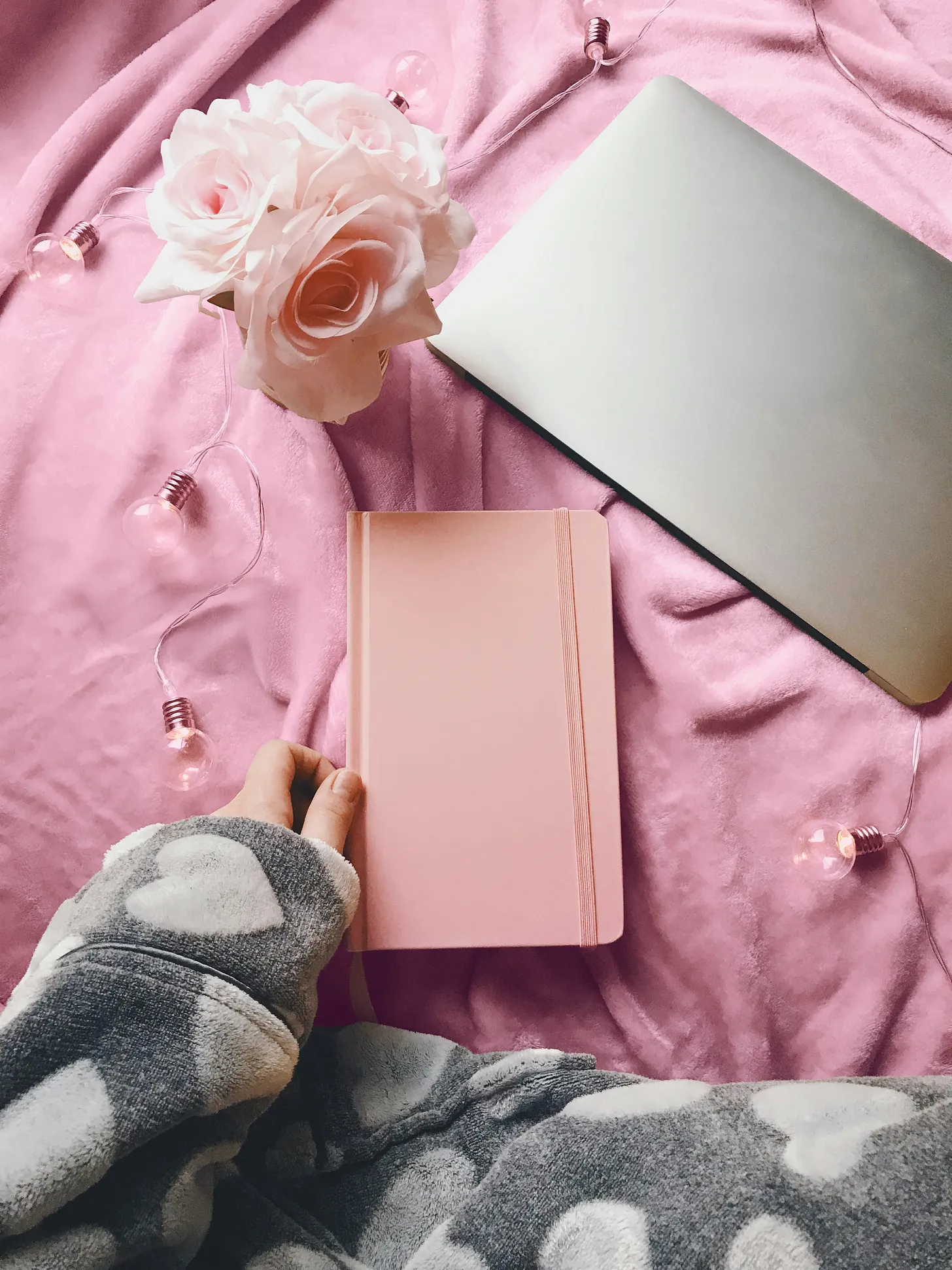 Woman in gray heart pajamas holds a peach notebook on a pink blanket, a laptop, and peach flowers.