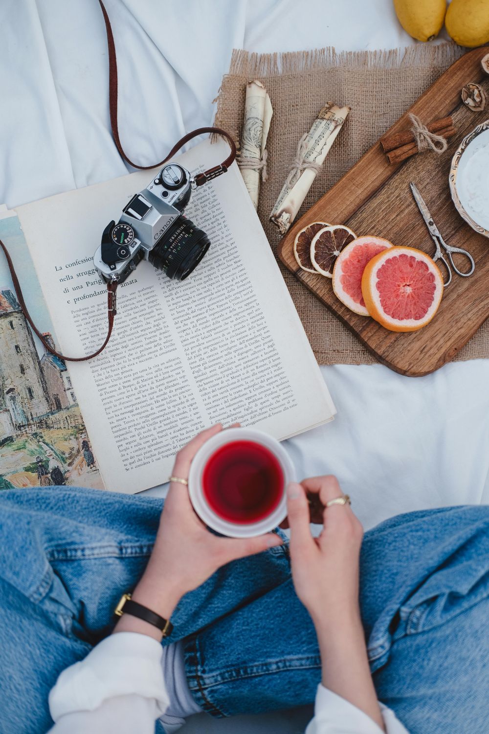 Woman outside drinking tea, surrounded by mandarins, book, camera, and cozy decor.