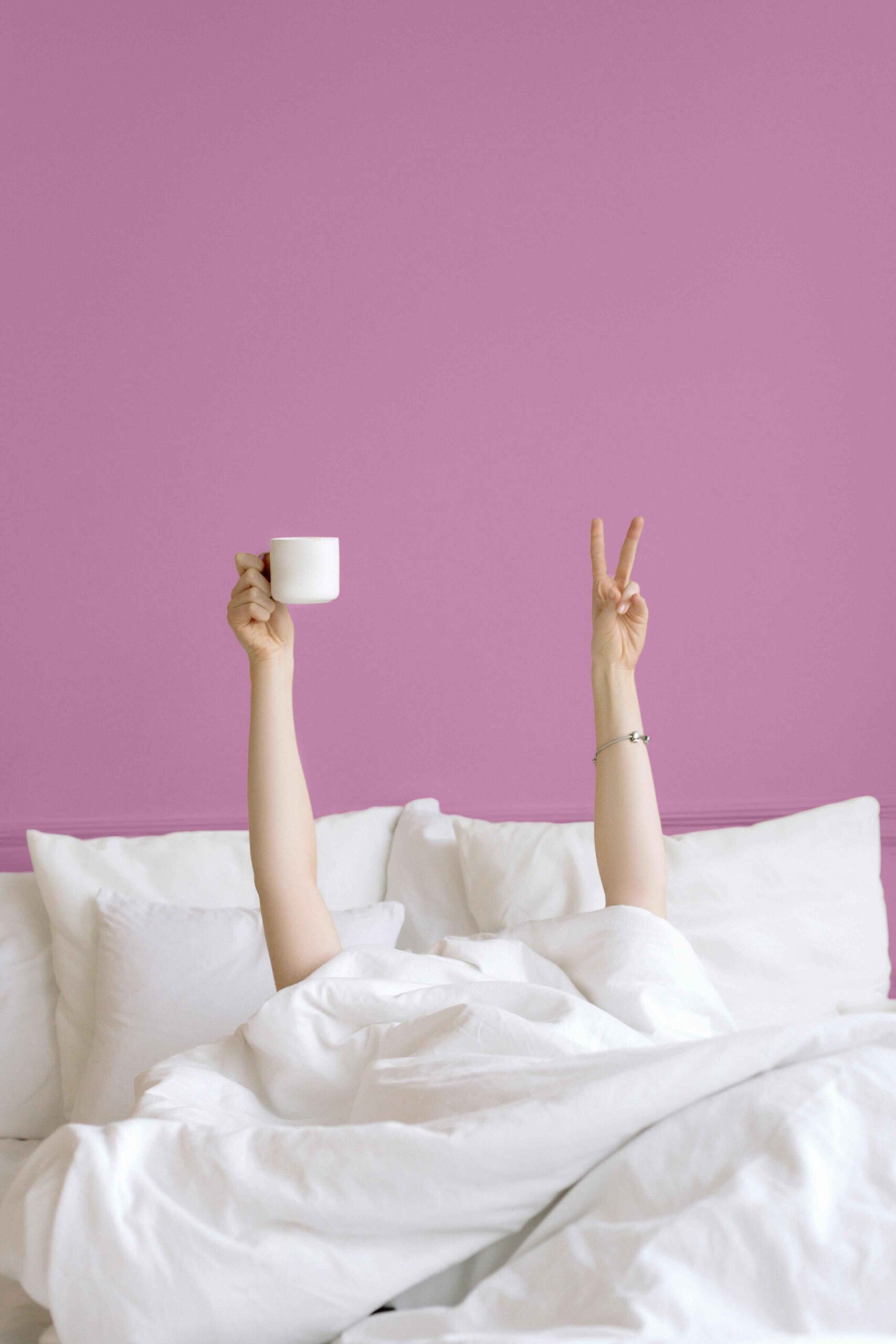Hands holding a white cup and making funny gestures while lying in bed, posed against a pink wall.