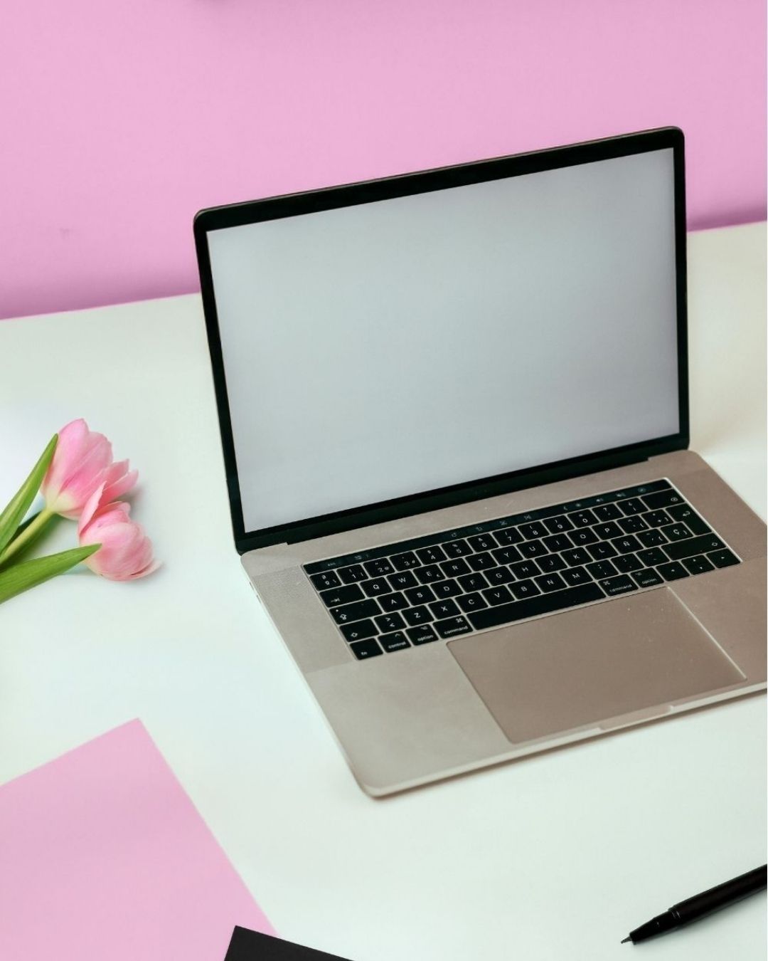 White desk with a laptop, pink flowers, pink and black papers, black ballpoint pen, and a pink wall.