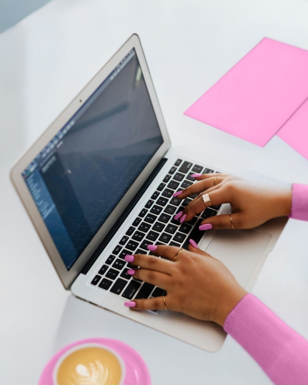 Woman in pink turtleneck, holding coffee, working on laptop with pink papers and creating templates.
