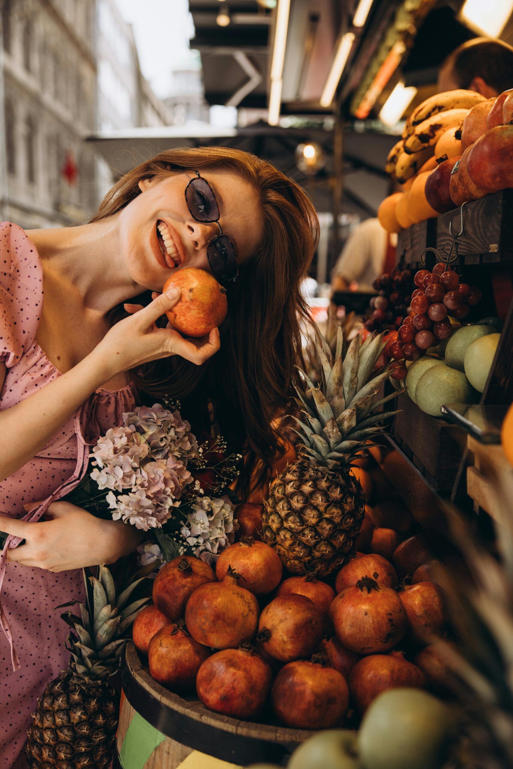 A woman with long red hair and sunglasses stands at an outdoor fruit stand. She is wearing a pink dress and smiling while holding a pomegranate above her head and a bouquet of flowers in her other hand. In front of her, the stand is filled with various fruits, including pineapples and a large pile of pomegranates.