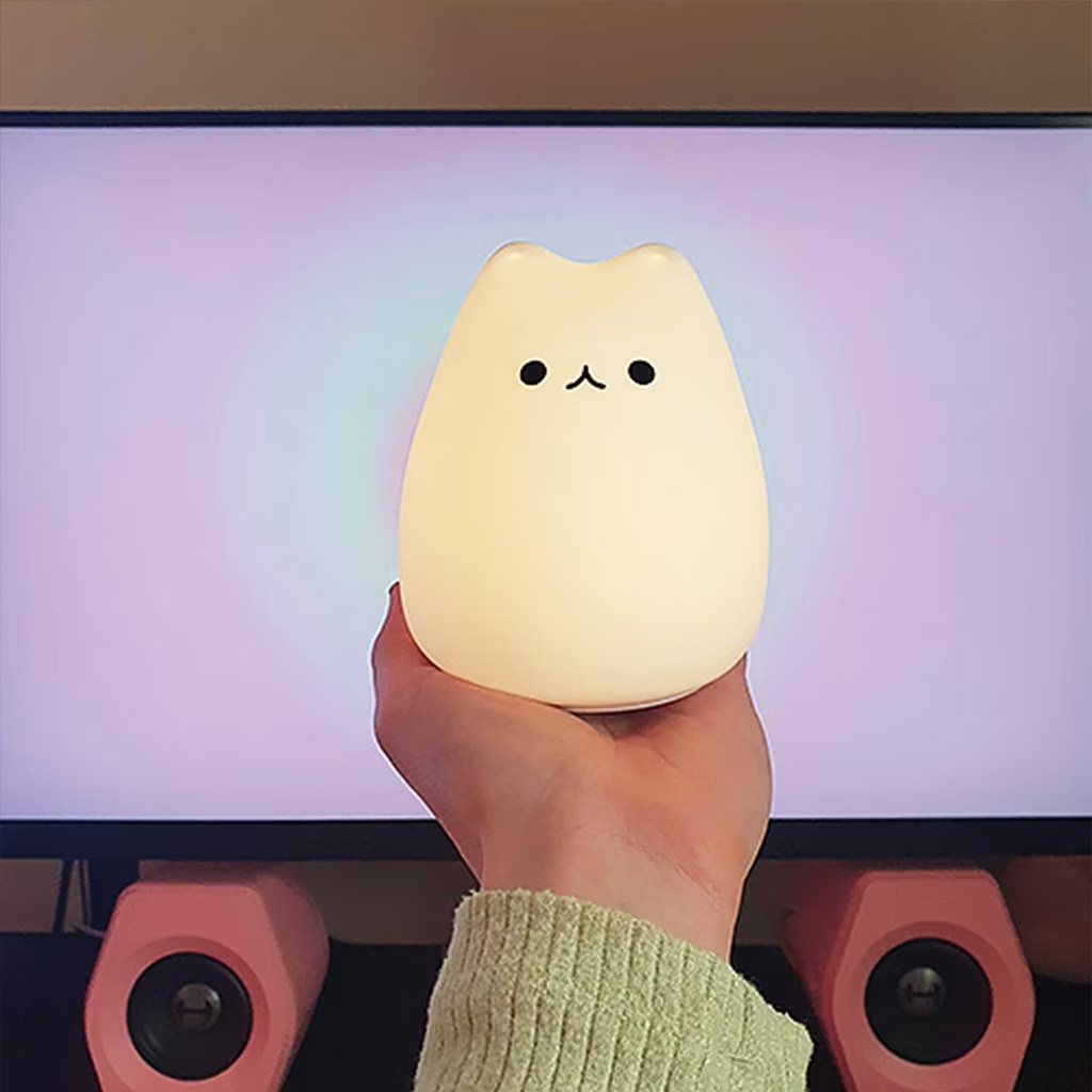 Person’s hand holding The Cat Lamp, a squishy silicone night light with a warm glow and a simple face. This cute lamp, a favorite for cozy office and desk aesthetics, appears on the content creator essentials list.