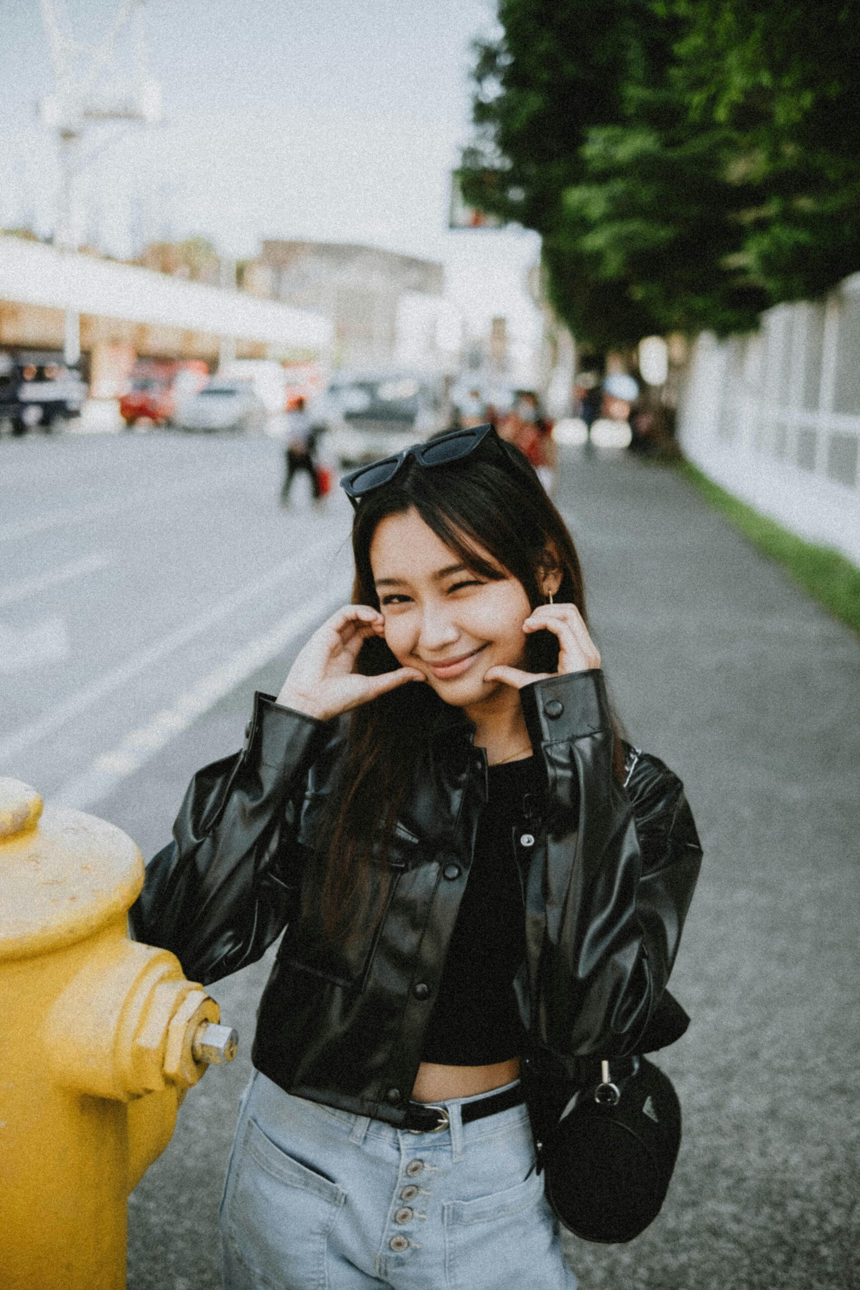 A young woman in a black leather jacket and jeans winking and making a playful face, leaning against a yellow fire hydrant on an urban street.