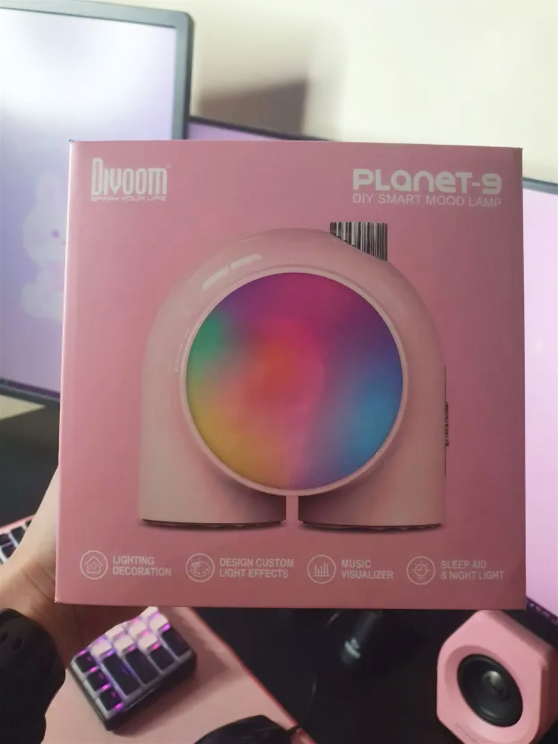 Hand presenting the pink box of the Divoom PLANET-9 DIY Smart Mood Lamp, one of the content creator must-haves. The box shows a rounded, pink, arc-shaped light with a rainbow gradient and highlights features like lighting decoration, custom light effects, music visualizer, and sleep aid night light.
