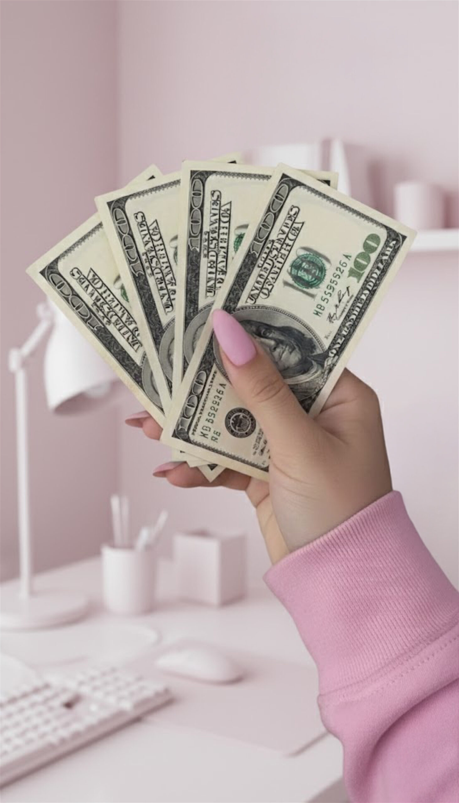 Hand with pink manicure and sleeve holding a fan of five $100 US Dollar bills against a blurry, minimalist background of a desk and wall in various shades of light pink.