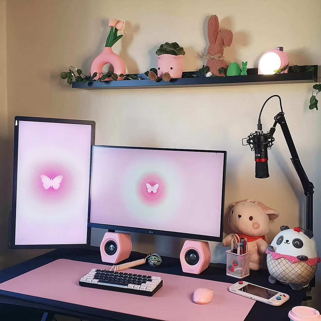 Brightly lit, pink aesthetic desk setup for a content creator, featuring two monitors with matching butterfly wallpapers, a white and pink mechanical keyboard, a pink mouse, pink speakers, and a black microphone on a boom arm. The floating shelf above has various plush toys and pink office decor, including the Divoom lamp mentioned in the blog's essentials list.