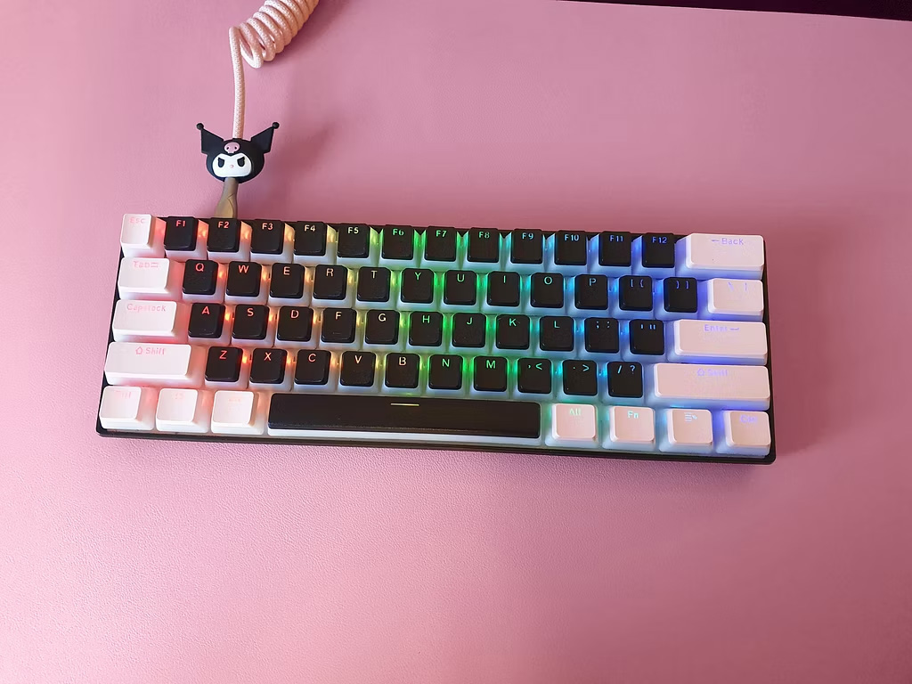 Close-up view of the customized RK61 Mechanical Keyboard with rainbow RGB lighting, black and white pudding keycaps, and a white coiled cable on a pink desk mat. This durable, aesthetic keyboard is a recommended essential for content creators.