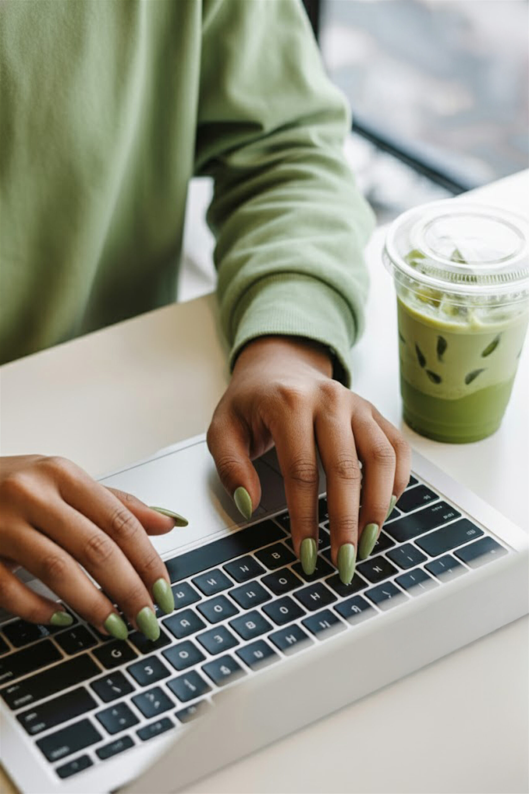 Hands with olive-green painted nails typing on a laptop, with a matching beverage nearby.