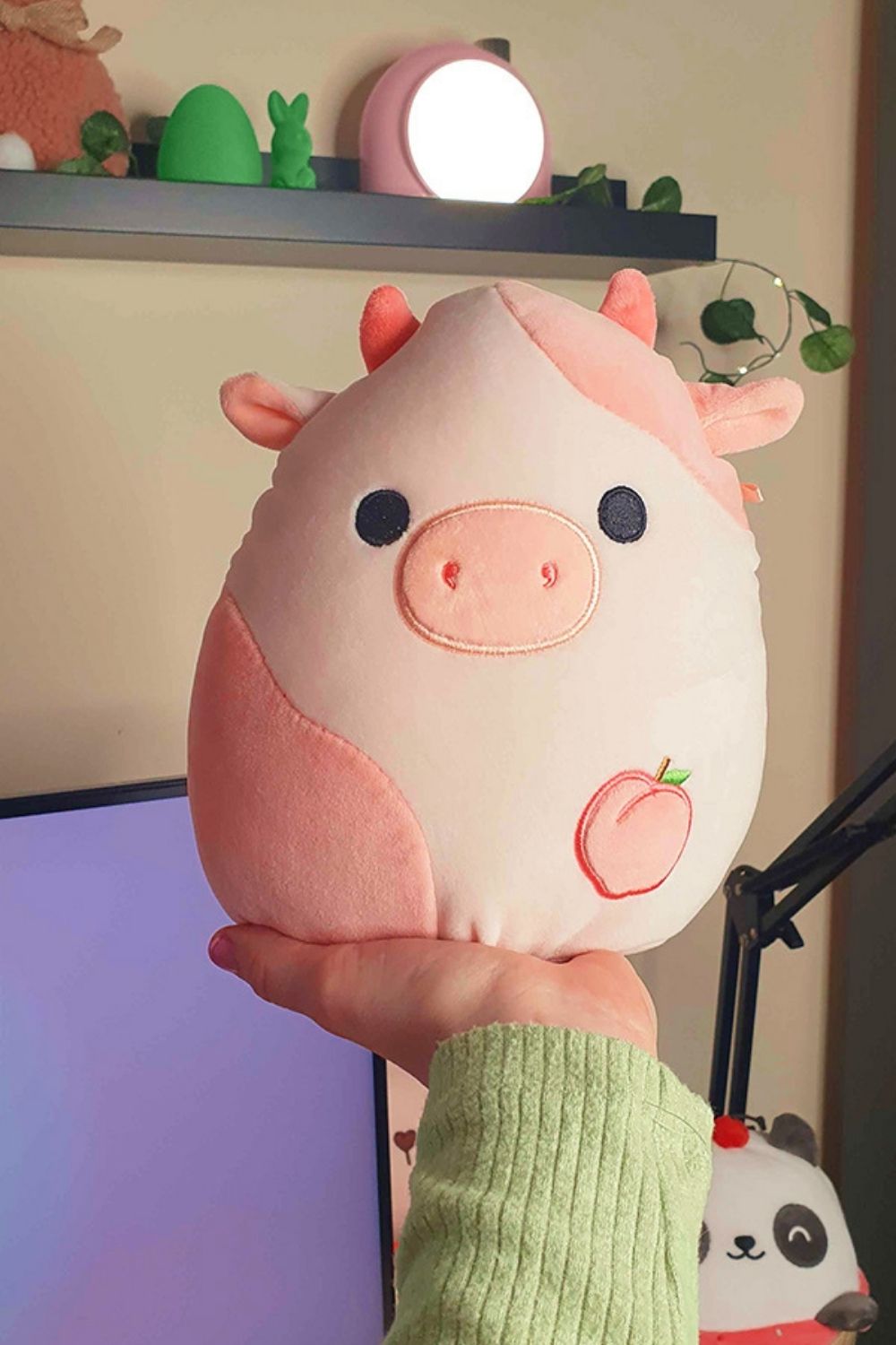 Large plush pink and white cow Squishmallow with a peach embroidered on its belly being held, one of the cozy content creator must-haves for an aesthetic home office setup, shown alongside other cute decor like a Divoom lamp in the background.