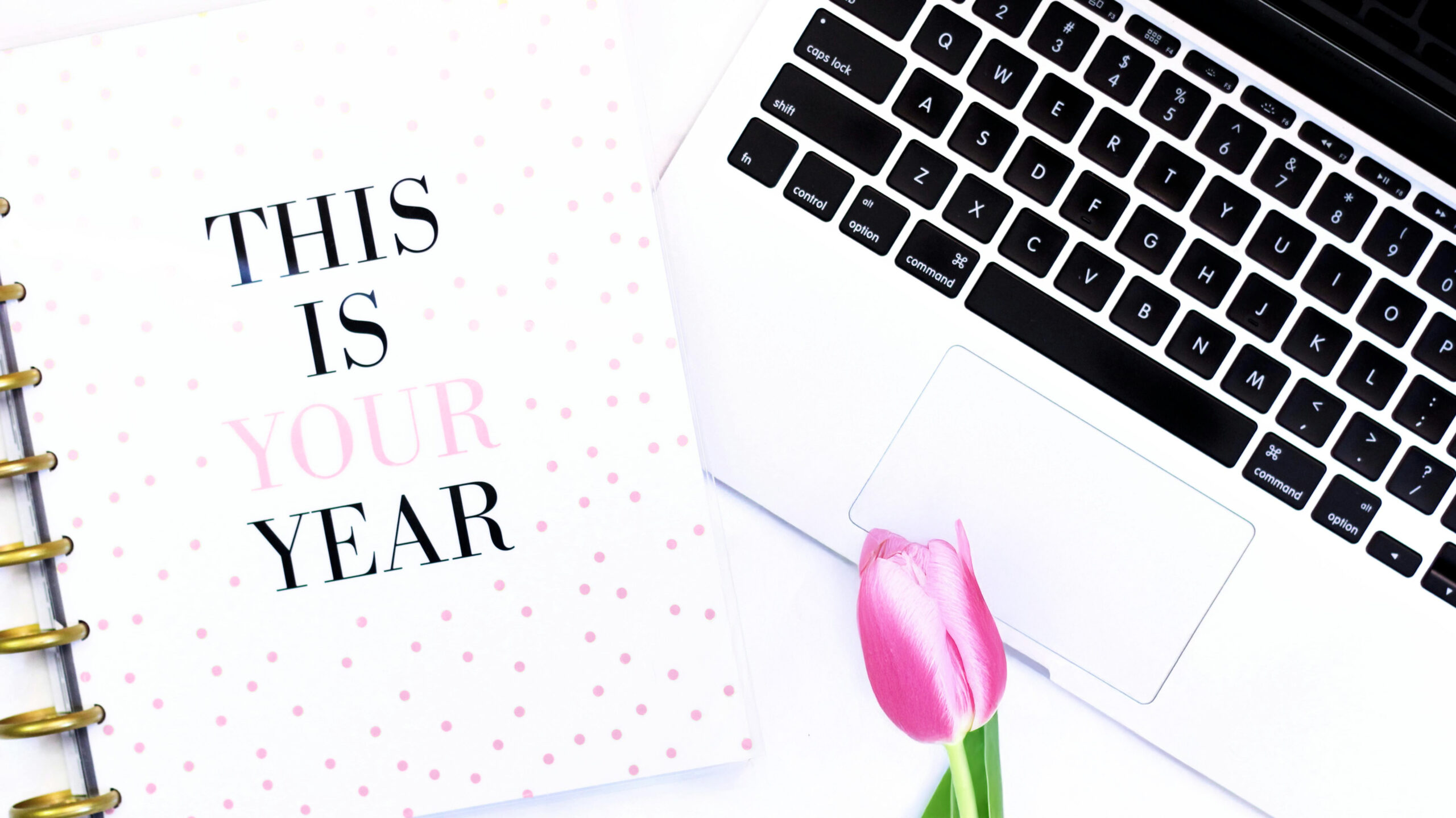 An overhead close-up flat lay shot of a white and gold spiral-bound notebook on the left with the text "THIS IS YOUR YEAR" (with "YOUR" in pink) on a white cover with pink polka dots, next to the keyboard of a silver laptop on the right. A single pink tulip rests on the white surface between the notebook and the laptop.