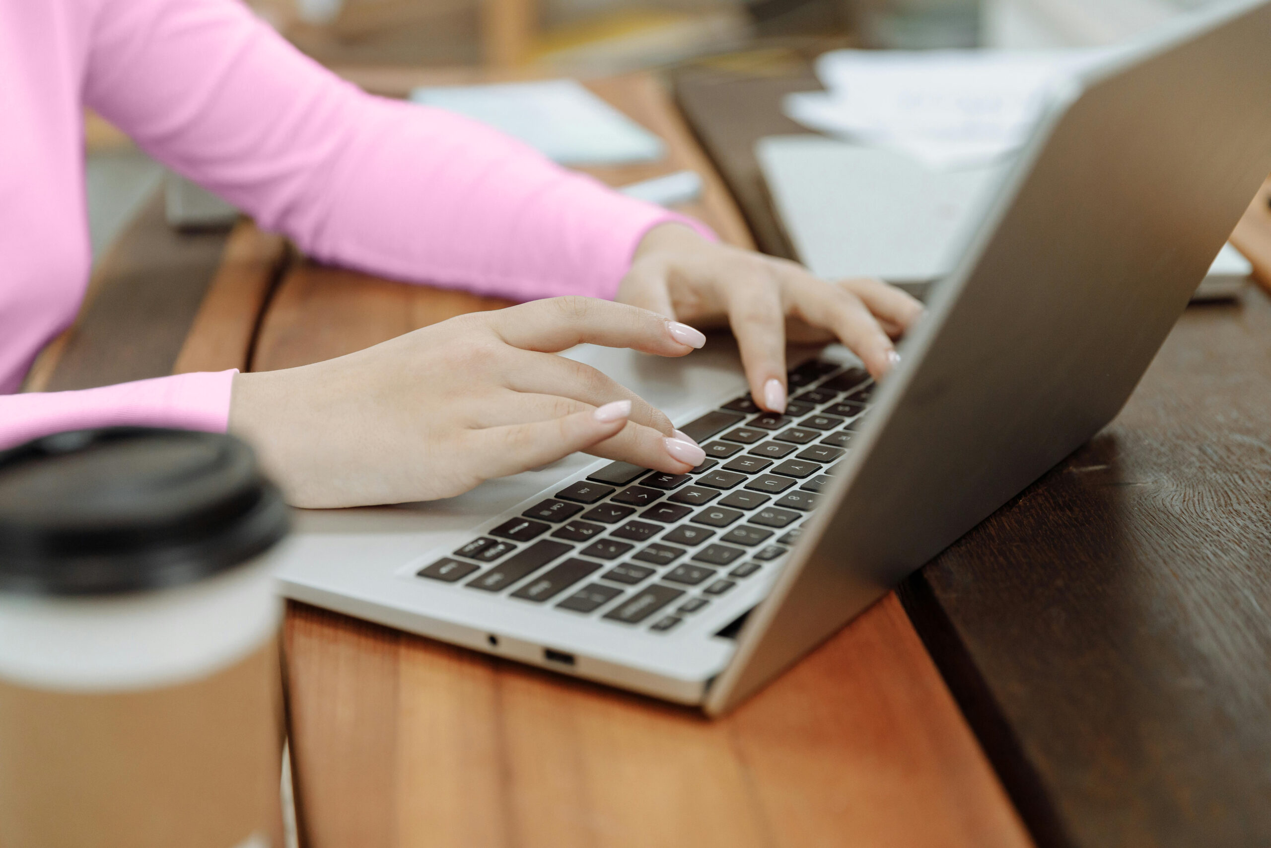 Close-up of a person wearing a pink long-sleeve T-shirt typing on a laptop keyboard at a wooden table with a to-go coffee cup, visually representing the creation and development process for one of the 100 digital product ideas.
