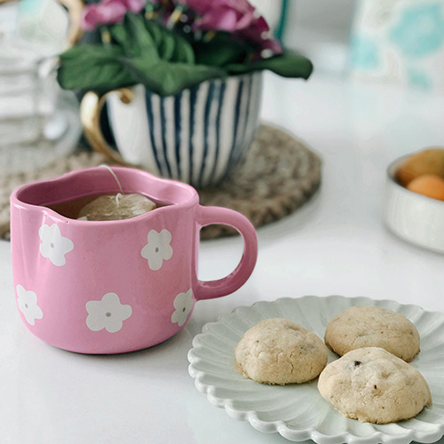 A pink mug with white flower patterns sits on a white table, a tea bag steeping inside. Next to it, a scallop-edged white plate holds three round cookies. In the blurred background, a blue and white striped teacup with a gold handle contains a plant with purple flowers and green leaves, and a bowl of fruit is visible.
