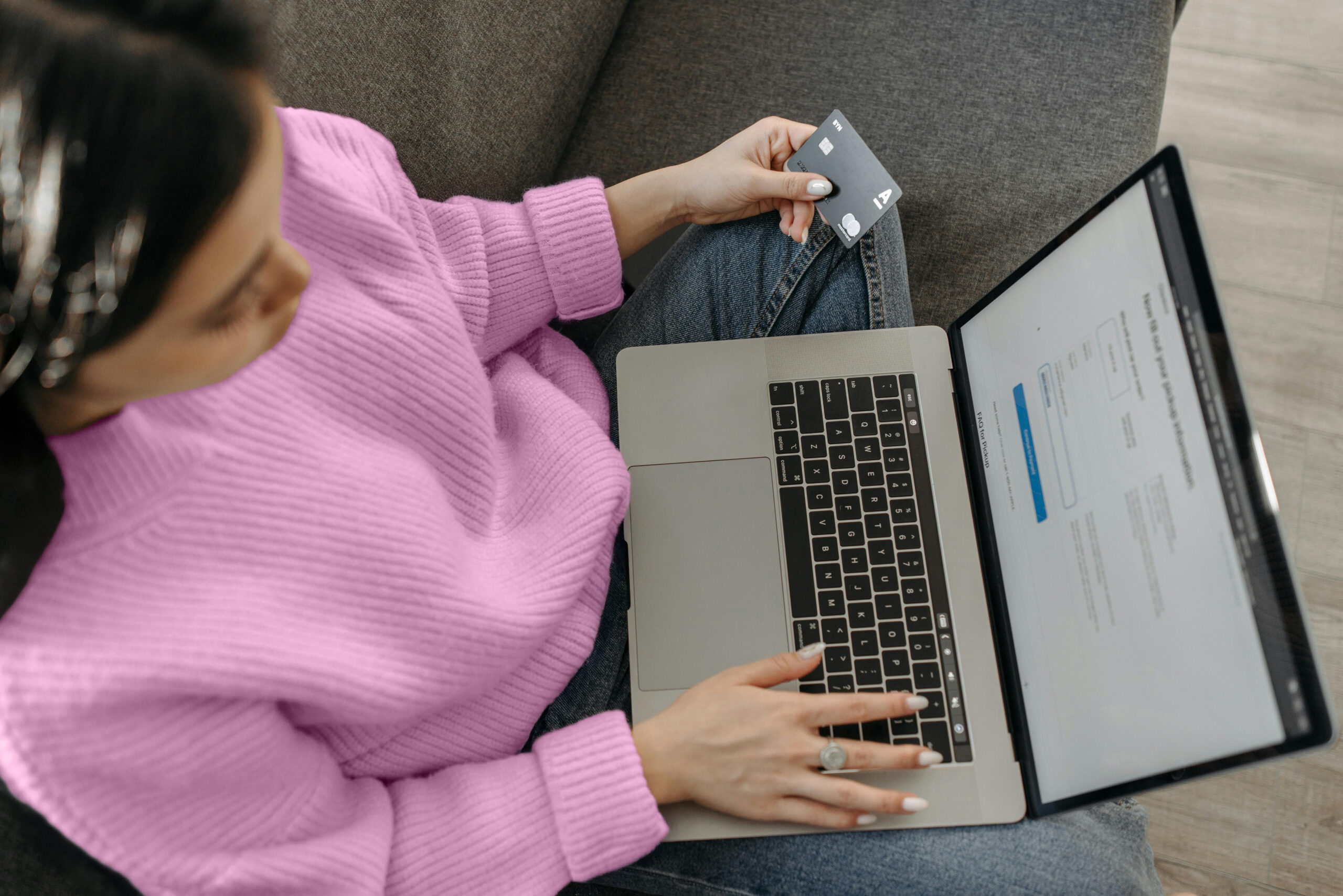 An overhead shot of a person sitting on a gray couch, wearing a pink sweater and jeans, using a silver laptop. They are holding a dark-colored credit card in their left hand while their right hand rests on the laptop's keyboard. The laptop screen displays what appears to be an online form or checkout page.