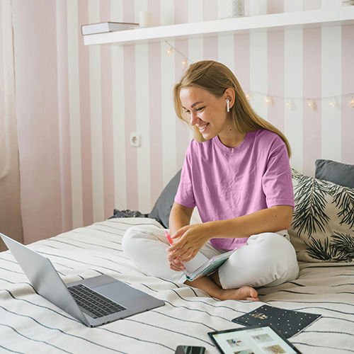 A young woman with blonde hair, wearing a baby pink t-shirt and white pants, is sitting cross-legged on a bed, smiling at a silver laptop. She has white wireless earbuds in her ears and is holding a small notebook and pen. The background features a wall with vertical pink and white stripes and a white floating shelf decorated with string lights. A tablet and a smartphone are visible on the striped bed cover in front of her.