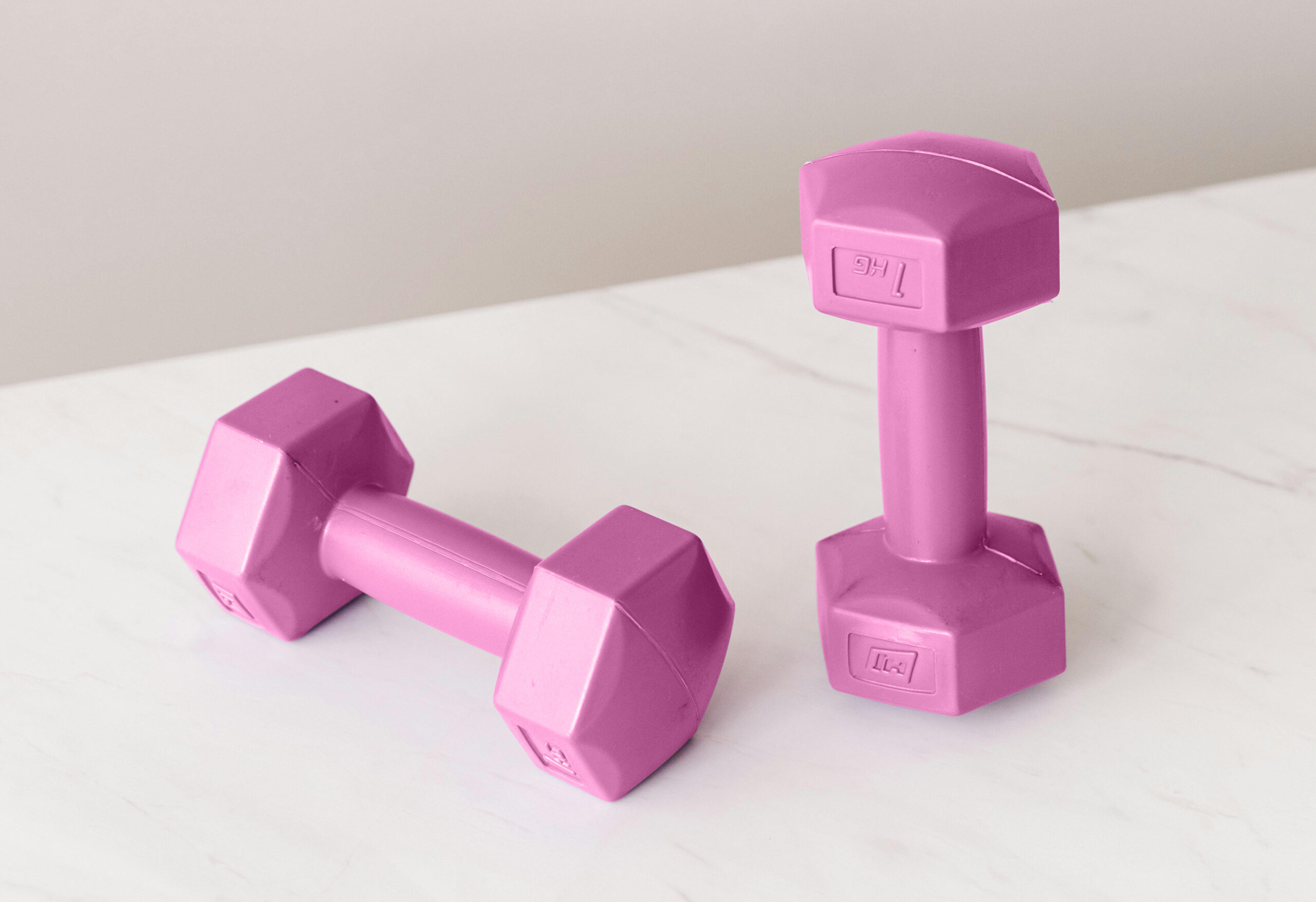 A pair of small, pink hexagonal dumbbells (hand weights) resting on a white marble surface, an image visually representing the wellness niche and inspiring one of the 100 digital product ideas for fitness or health.