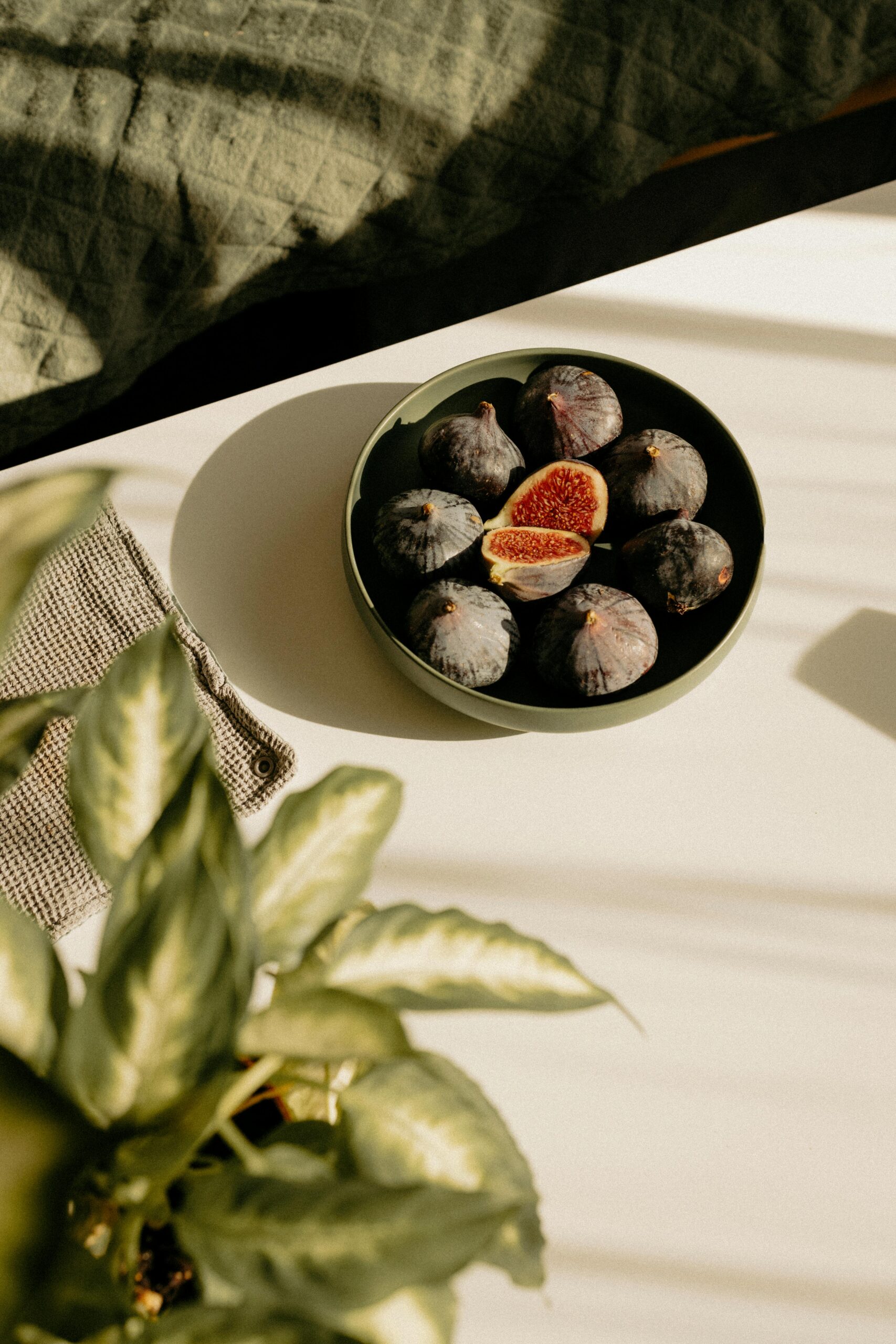 A bowl of dark purple figs, one cut open, sits on a sunlit white table. Soft green plant leaves and a waffle-textured cloth add a natural, serene vibe.