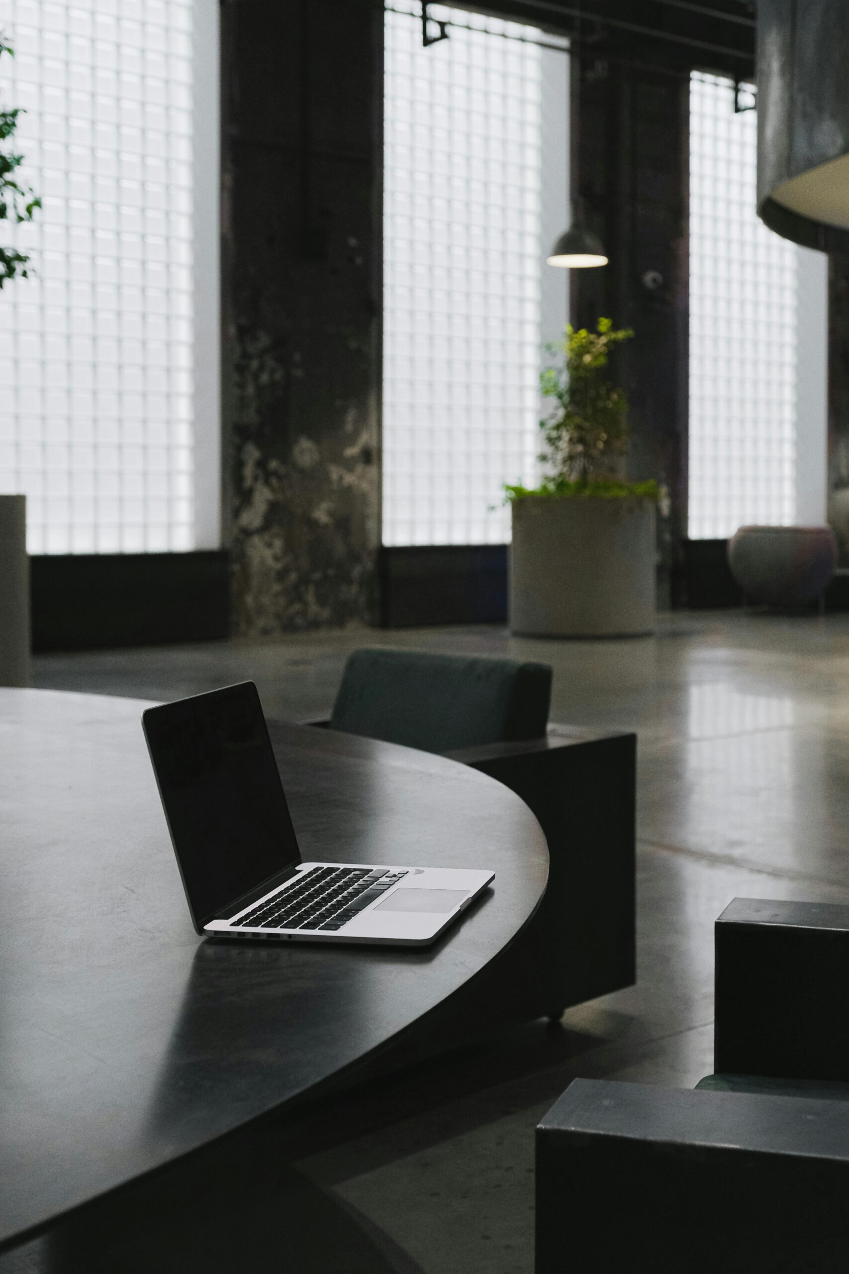 Sleek laptop open on a modern black table in an industrial-style room with large glass block windows, concrete floors, and lush potted plants.
