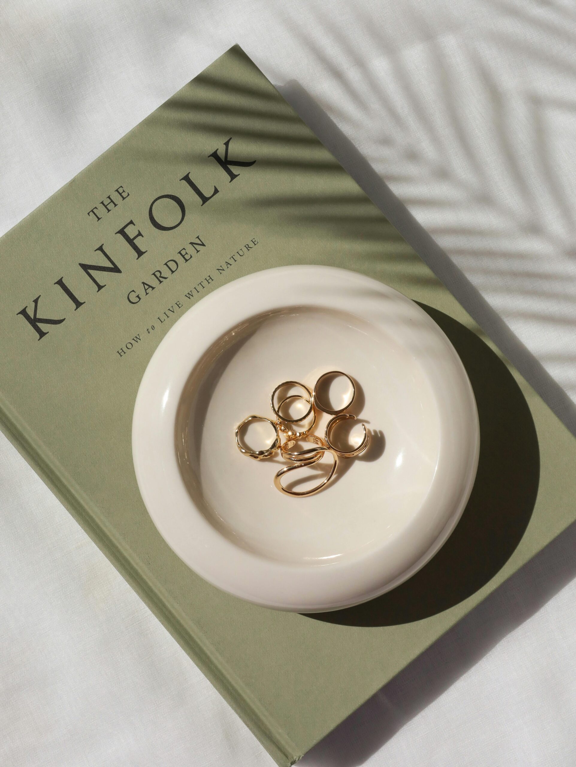 A serene image of gold rings in a white ceramic dish on a green book titled "The Kinfolk Garden." Soft shadows and a calm, minimalist tone.