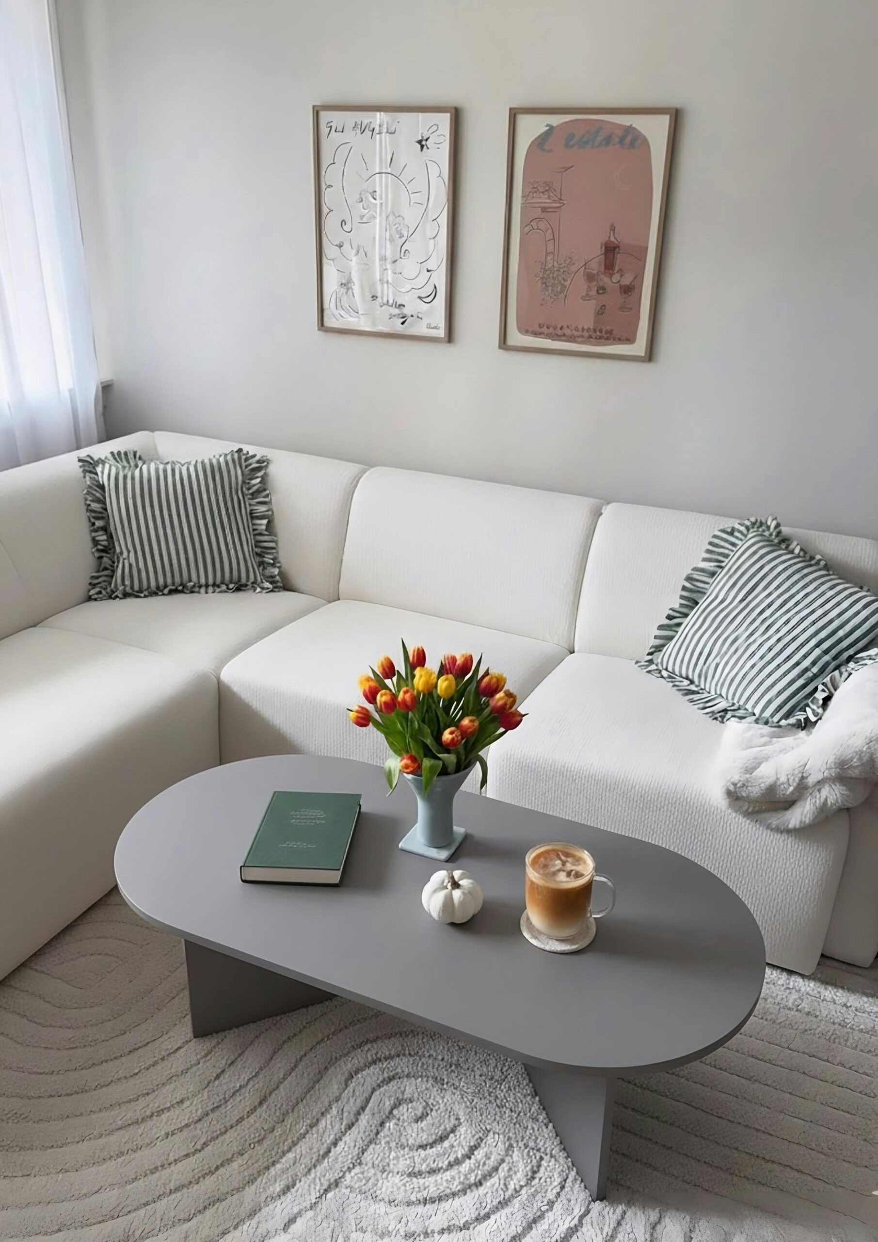 Cozy living room with a white sectional sofa, striped pillows, and a gray table. Tulips in a vase, book, small pumpkin, and latte on the table. Relaxed ambiance.