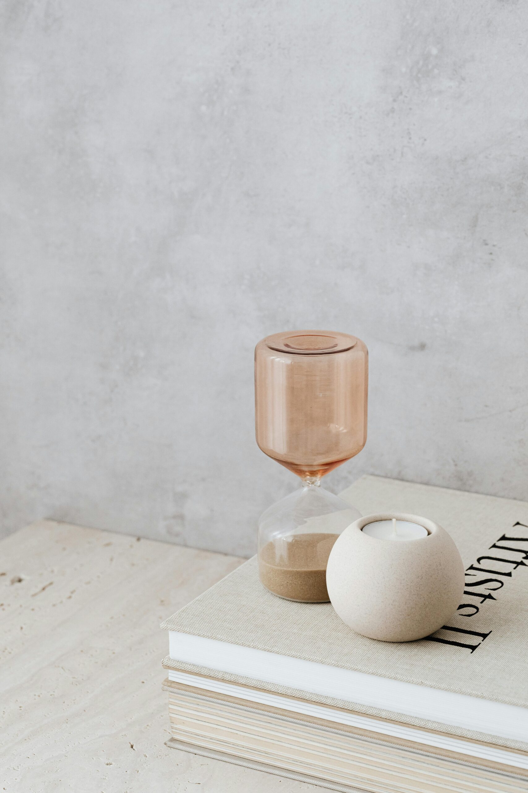 A minimalist scene with a brown hourglass and a round beige candle on stacked books, set against a gray textured wall. Calm, neutral tones prevail.