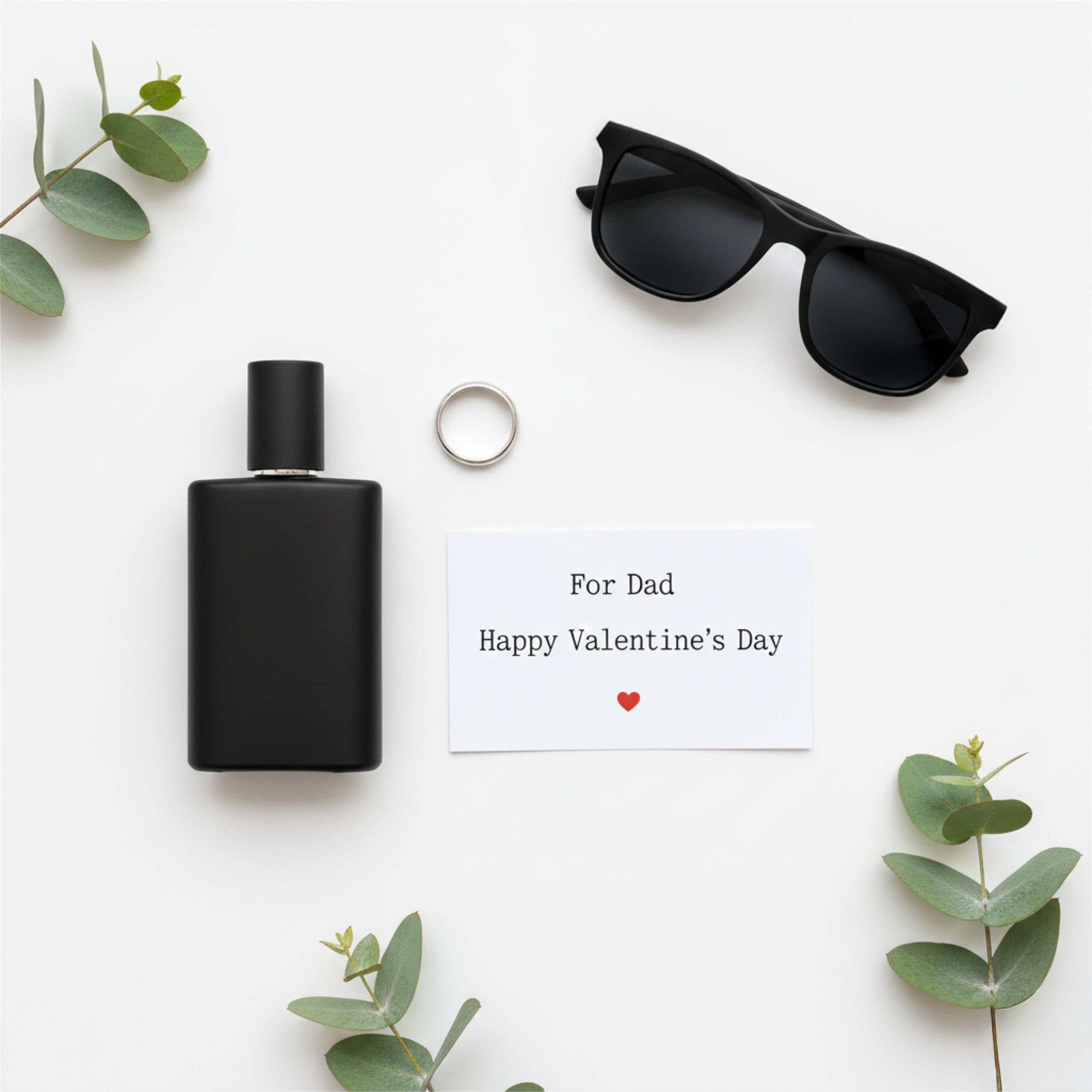 Flat lay of a black perfume bottle, sunglasses, a ring, and a card reading "For Dad, Happy Valentine's Day" with a red heart. Greenery accents.
