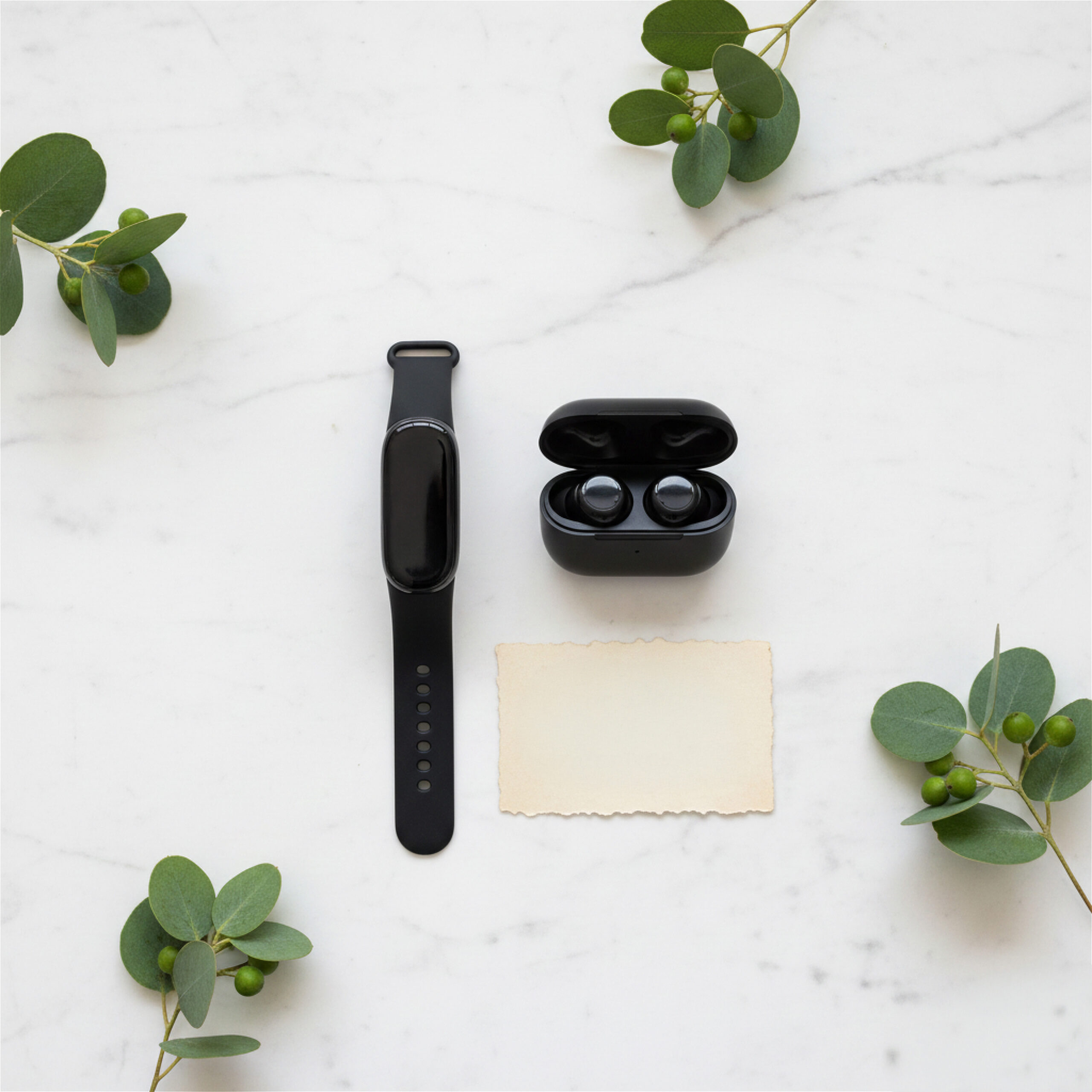 Black smartwatch and earbuds on a white marble surface, surrounded by green leaves. A blank cream card sits nearby, creating a fresh, minimalist tone.