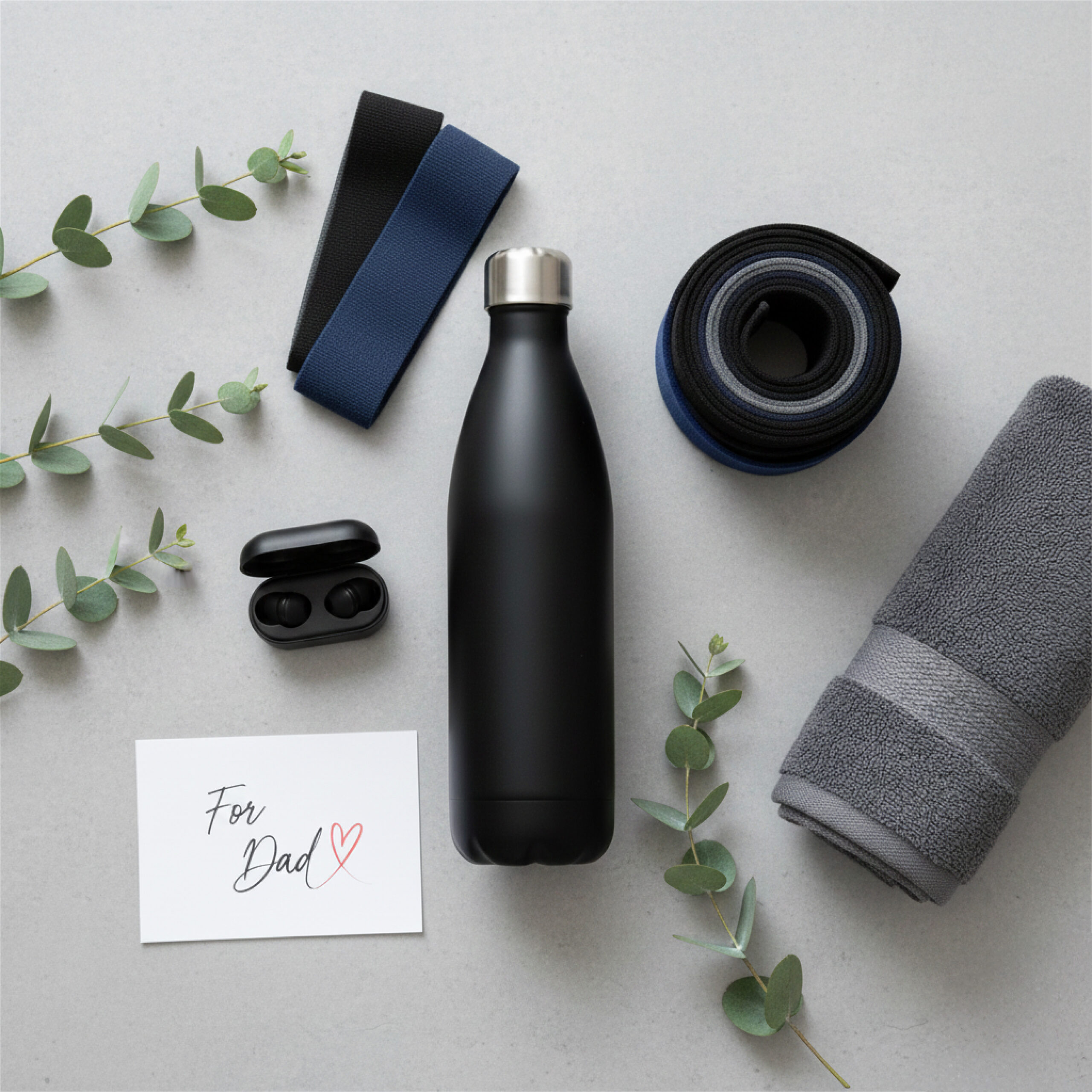 Flat lay with a black water bottle, earbuds, resistance bands, a rolled towel, eucalyptus sprigs, and a card reading "For Dad" with a heart.