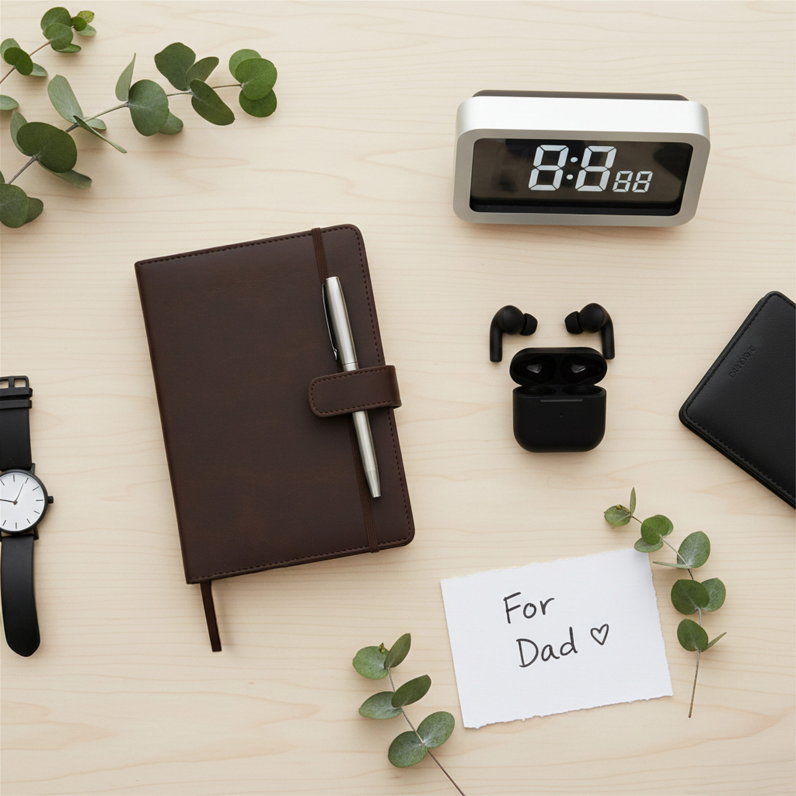 Desk arrangement with a modern alarm clock, black earbuds, a brown leather journal, watch, black wallet, and note reading "For Dad" with greenery.