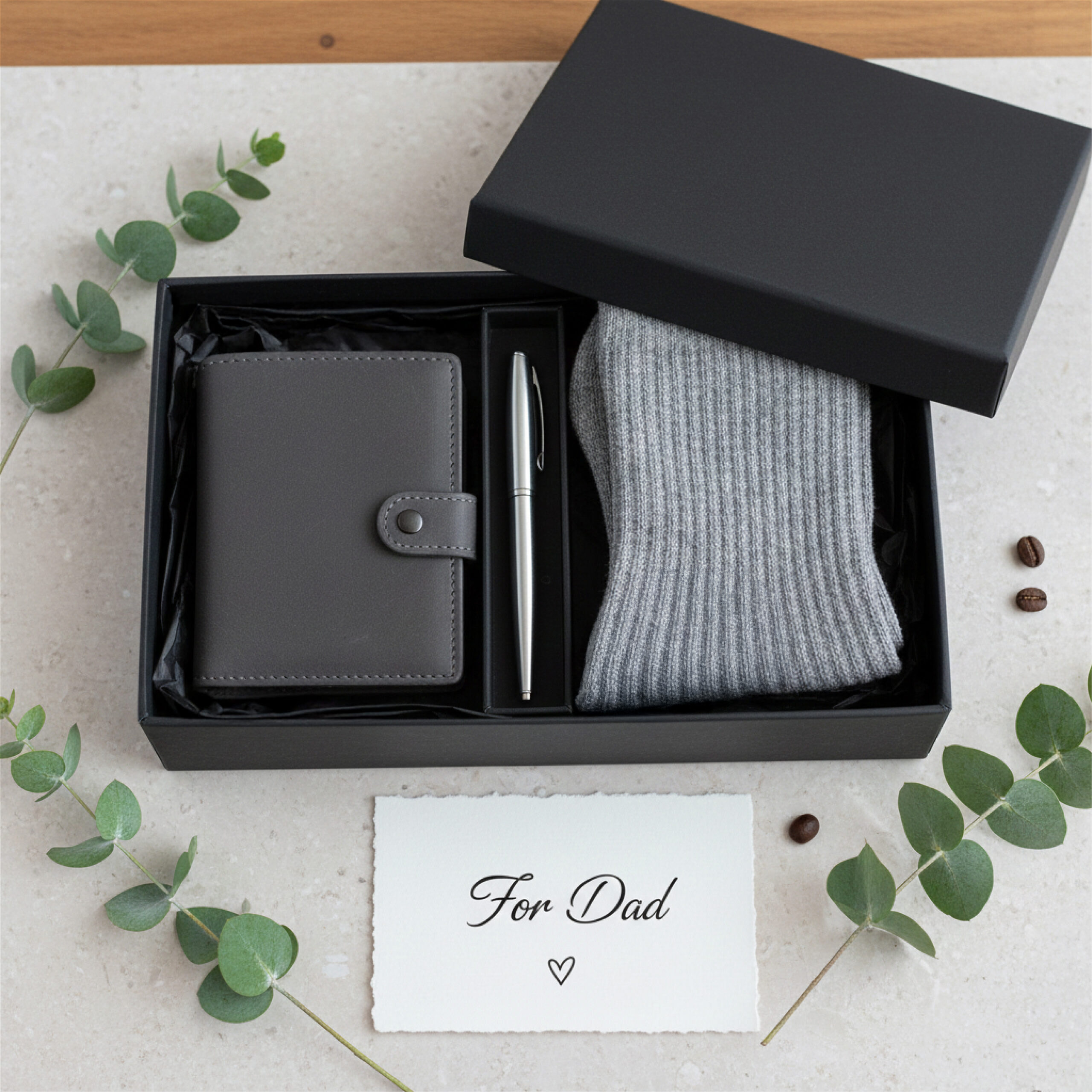 Gift box with a gray wallet, silver pen, and gray socks arranged neatly. A card reads "For Dad" on a marble surface, surrounded by eucalyptus.