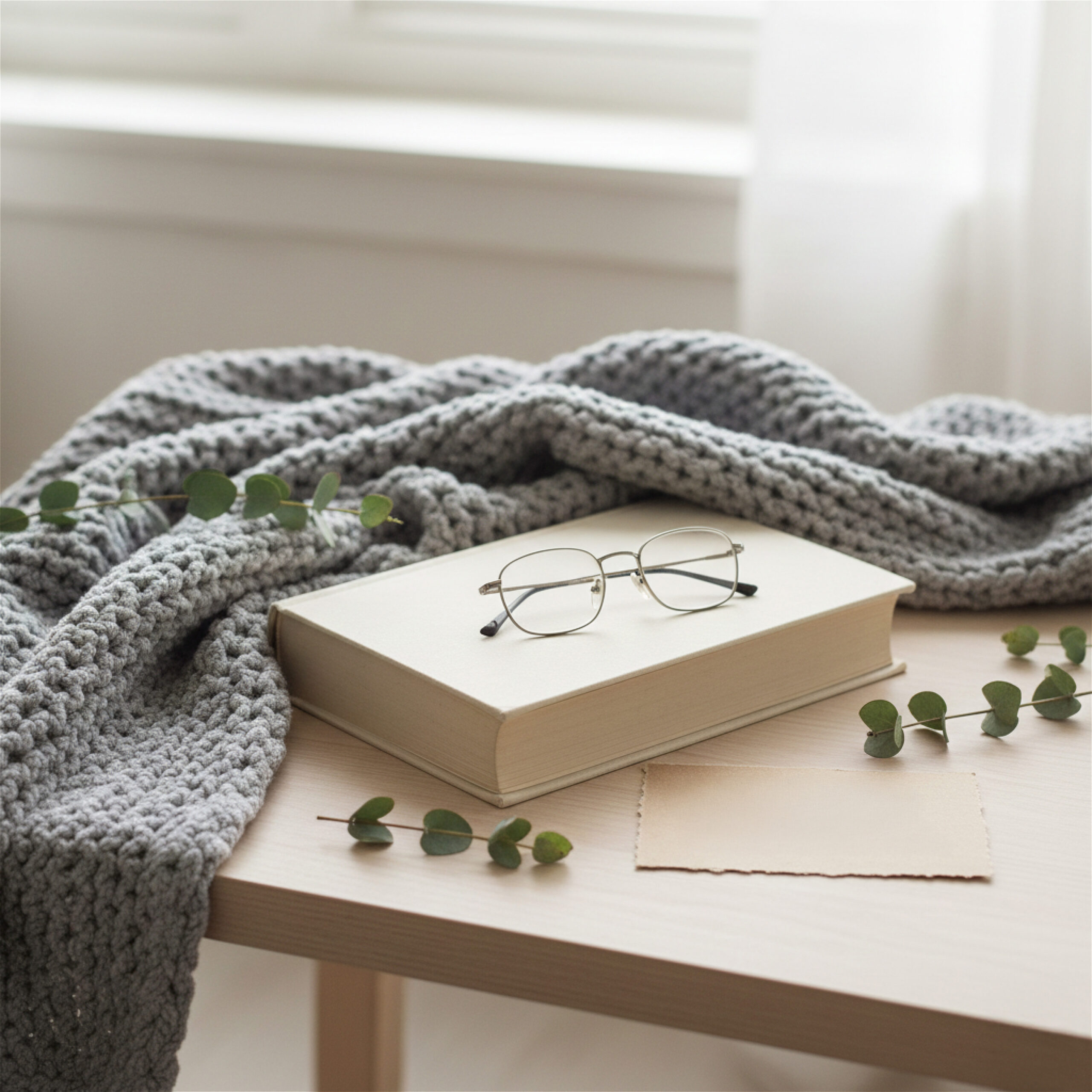 A cozy scene with glasses on a closed book, draped with a gray knit blanket. Green leaves and soft lighting evoke a calm and inviting atmosphere.
