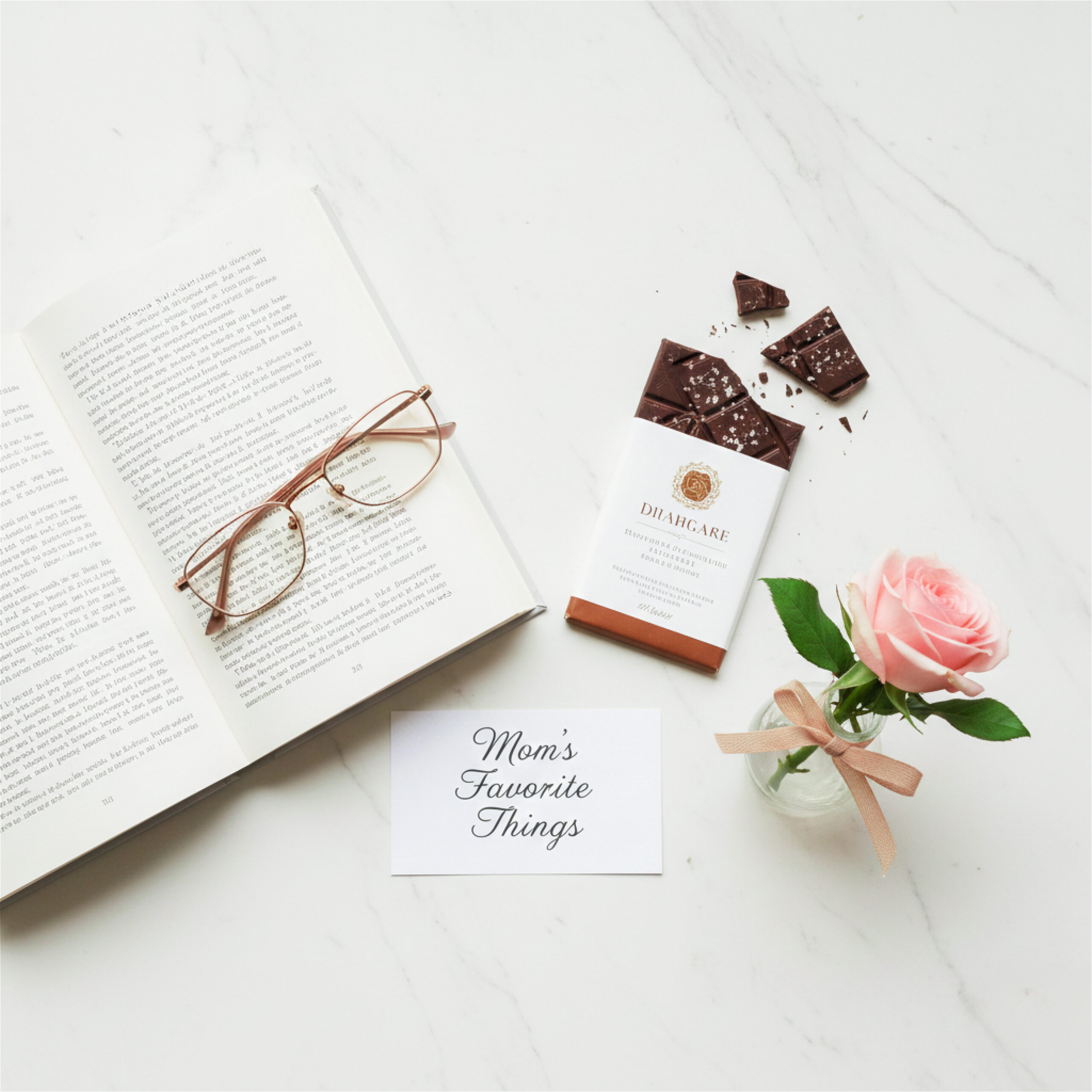An open book with reading glasses, a chocolate bar with broken pieces, a pink rose in a vase, and a card reading "Mom's Favorite Things" on a white surface. This thoughtful arrangement offers lovely ideas for Valentine's Day presents for mom.