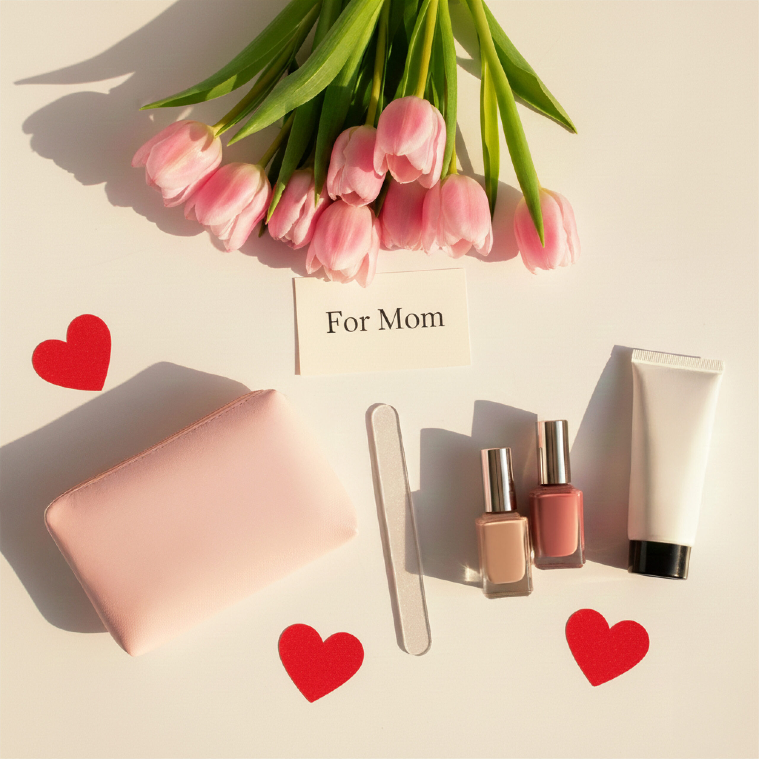 Pink tulips and a "For Mom" card rest above cosmetics, a nail file, a pink pouch, and red hearts on a white surface, conveying love and care.