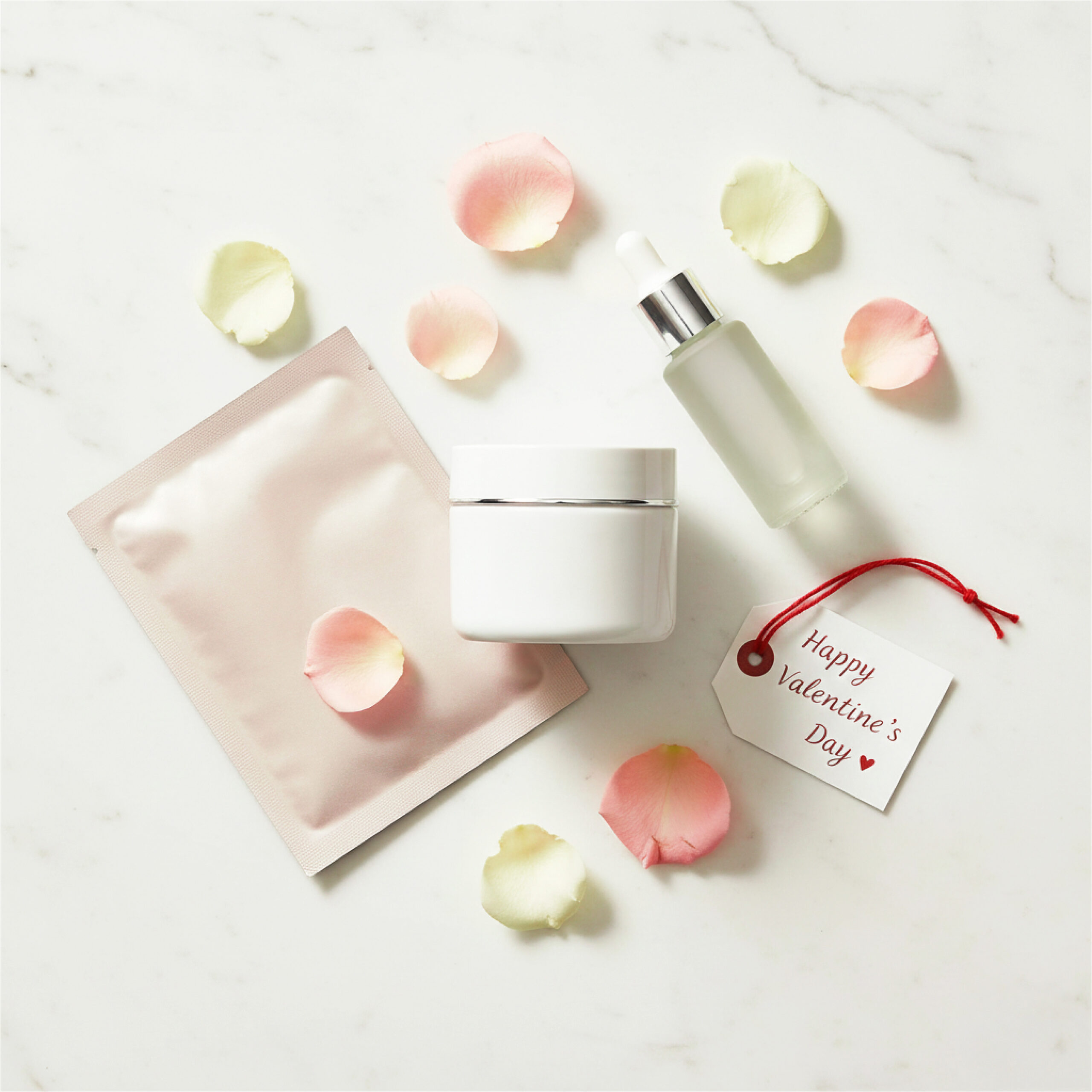 Skincare items on marble: a cream jar, serum bottle, and sheet mask surrounded by pastel rose petals. A tag reads "Happy Valentine's Day." These luxurious items make excellent Valentine's Day presents for mom.
