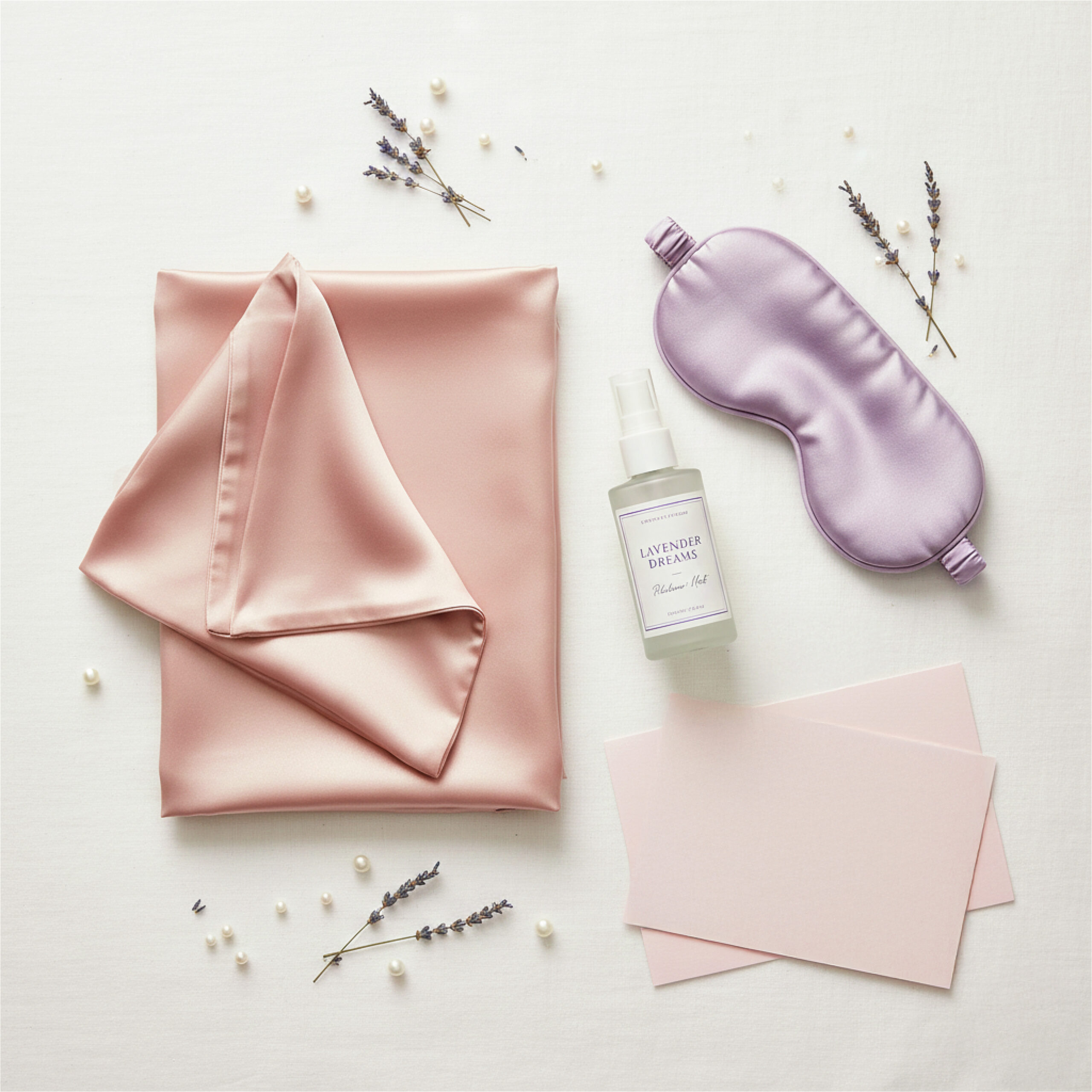 Silky pink fabric with a folded edge, lavender sleep mask, and spray labeled "Lavender Dreams" on cream surface, surrounded by lavender sprigs and pearls. Calm and serene.