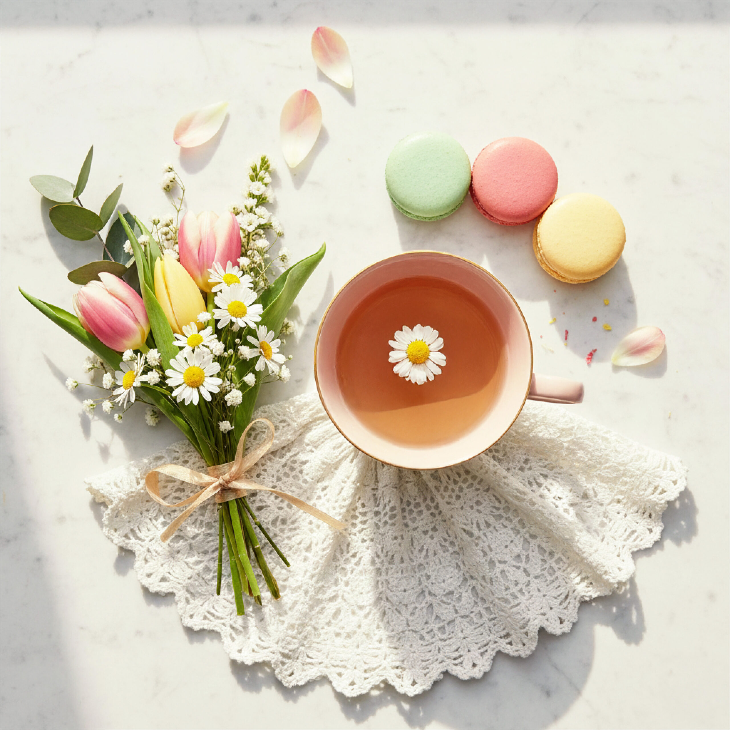 A tea cup with chamomile on lace, surrounded by tulips, daisies, and three colorful macarons on a marble surface, creating a fresh, serene mood. This delightful scene provides inspiration for thoughtful Valentine's Day presents for mom.