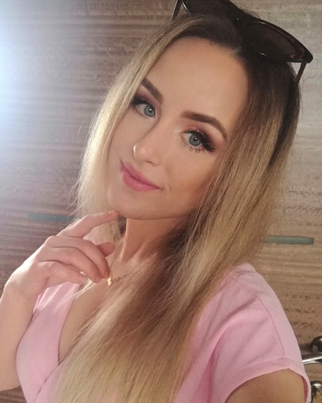 A person with long blond hair, wearing sunglasses on their head and a pink top, poses for a selfie with a smile, in front of a wooden wall.