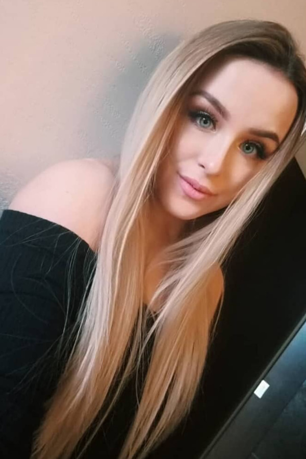 A woman with long, straight blonde hair and bright eyes smiles slightly, wearing a black off-shoulder top. The background is softly lit, evoking a warm, relaxed feeling.