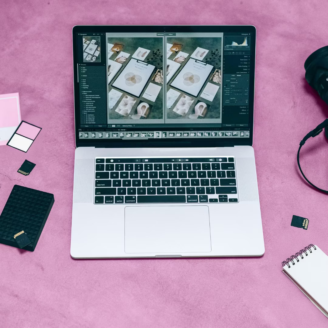 A laptop displaying photo editing software on a pink surface surrounded by a camera, memory cards, notepad, and color swatches, conveying creativity.