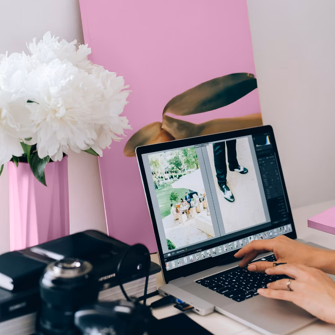 A person edits a wedding photo on a laptop in a bright workspace with a pink wall, white flowers, and a camera. The scene conveys creativity and focus.