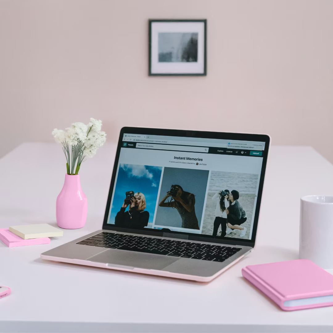 A laptop on a white desk displays a photography website. Nearby are a pink vase with flowers, pink notebooks, sticky notes, and a white mug.