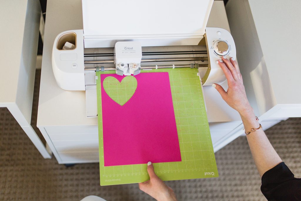 A person's hands operating a white Cricut cutting machine to cut out a heart shape from bright pink cardstock loaded on a green cutting mat.