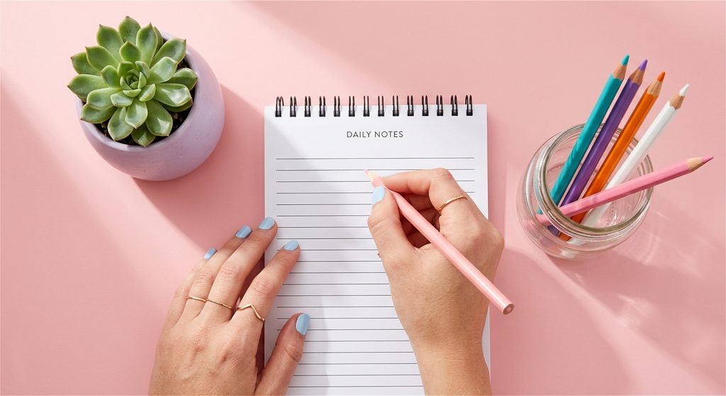 Top-down view of hands writing on a 'Daily Notes' spiral notepad with a pink pencil on a pastel pink background, alongside a green potted succulent, colored pencils in a jar, and teacher SVG project planning materials.